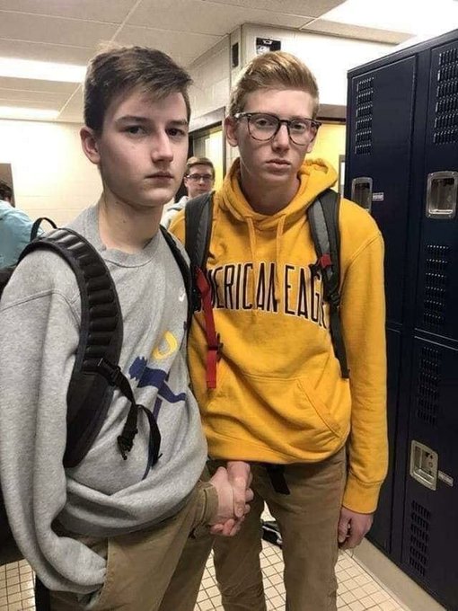 Two teen boys stand by school lockers, both wearing backpacks; one sports a gray sweatshirt with a horse graphic while the other wears a yellow "American Eagle" hoodie and glasses, looking unamused. Their deadpan expressions perfectly match the post's joke about "$INJ & $4.99" capturing a moment where absolutely nothing noteworthy is happening—just two kids existing.