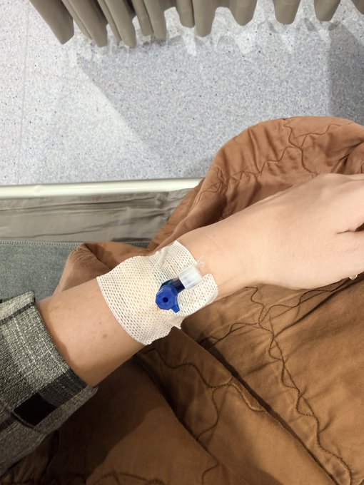 A close-up of a person's arm with an IV cannula secured by white gauze, resting on a brown blanket.
