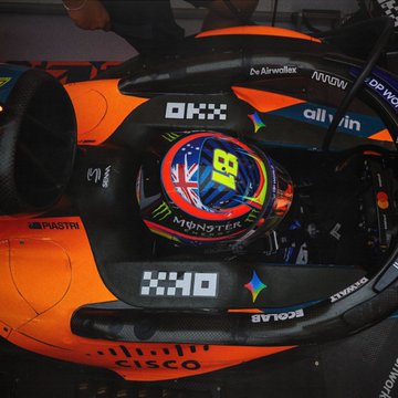 A McLaren Formula 1 driver's helmet featuring the number 81 sits in the cockpit of the orange Papaya livery car, surrounded by visible sponsor logos including OKX, Monster Energy, Dell, and Richard Mille. The image showcases the "new money" aesthetic of modern F1 sponsorship branding, with the driver's helmet prominently displayed against the vibrant orange chassis.