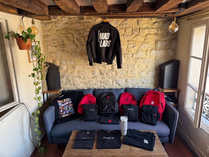A retail display or photograph showing a black "MAD LADS" bomber jacket hanging on a stone wall above a grey sofa lined with several red and black "Fuck it." branded backpacks and t-shirts.