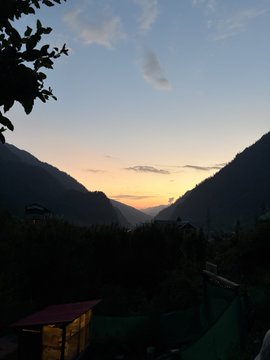 A sunset view over mountain silhouettes, with a warm orange glow on the horizon transitioning to a blue sky above, framed by foliage in the foreground.