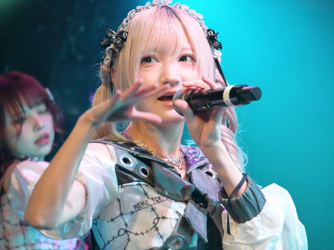 A J-pop/idol singer with pink hair and decorative bows performing on stage, holding a microphone and wearing a white and black outfit with 'MEME TOKYO' visible on the garment.