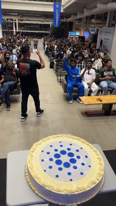 A scene from a Cardano event with a large crowd seated in a convention hall, a person taking a photo, and a large cake decorated with the Cardano logo in the foreground.