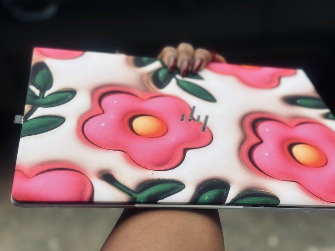 A photograph of a laptop lid or tablet featuring a sticker design with large pink flowers and green leaves on a white background, held up by a hand.