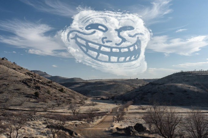 A dry, hilly landscape with a winding dirt road beneath a blue sky, featuring a large digitally-added cartoon face with a mischievous grin formed by the clouds.