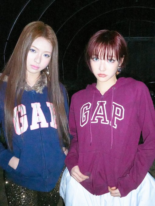 A photo of two young women wearing GAP hoodies—one navy blue with red lettering, one maroon with white lettering—styled with bold makeup and statement earrings.