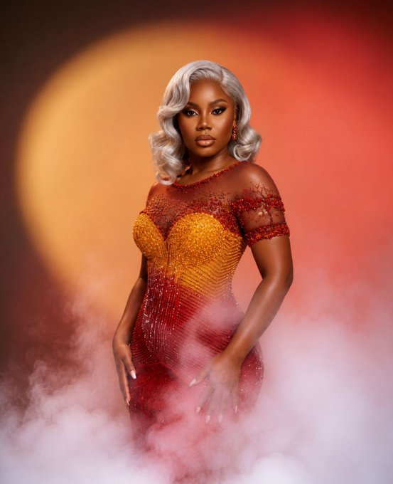 A glamorous portrait of a woman with silver hair wearing a sparkling red and orange dress, surrounded by pink smoke against a warm-toned background.