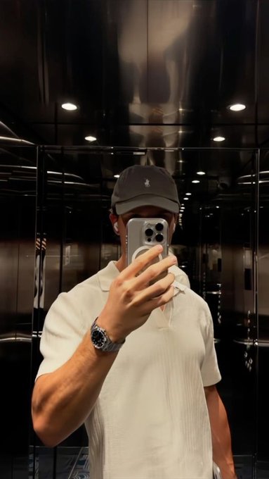 A man in a white polo shirt, dark cap, and luxury watch takes a mirror selfie in an elevator, holding a silver smartphone that obscures his face.