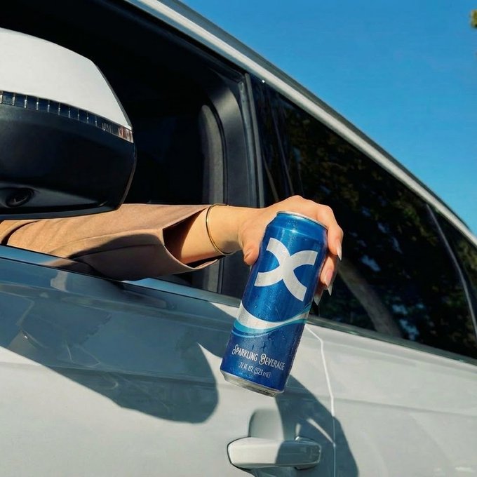 A hand extends from the driver's side window of a white car, holding up a blue can labeled "X SPARKLING BEVERAGE" with "ZERO SUGAR" visible. The image complements the post's theme of "a little fuel for the ride ahead" by playfully suggesting the beverage as alternative fuel for the journey.