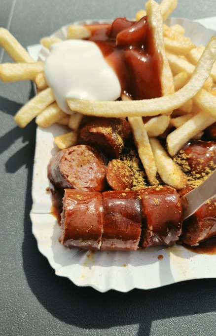 A paper tray holds sliced currywurst topped with sauce and curry powder, served alongside french fries and a dollop of mayonnaise.