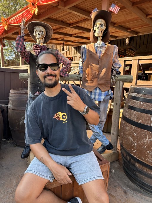 A man poses with a peace sign in front of two skeleton figures dressed in Western cowboy attire, one waving hello, both wearing cowboy hats and vests near wooden barrels. He's wearing a black t-shirt featuring a flame logo with "C" and what appears to be a "Rip Curl" or similar brand emblem, paired with light denim shorts. The scene captures a fun, themed photo-op moment perfect for a cheerful "GM fam" (good morning family) greeting to his social media followers.