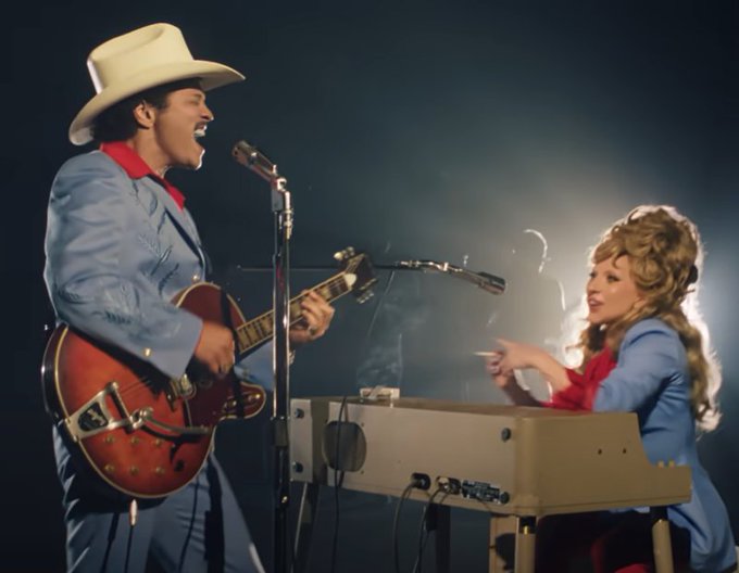 Two musicians perform on stage, with a man in a cowboy hat singing and playing a red electric guitar alongside a woman with voluminous blonde hair playing a keyboard.