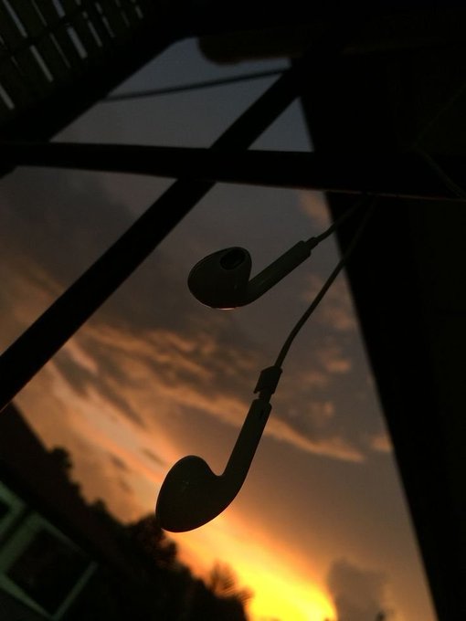 Silhouetted earbuds hang against a dramatic twilight sky with orange and dark clouds visible through a window frame.