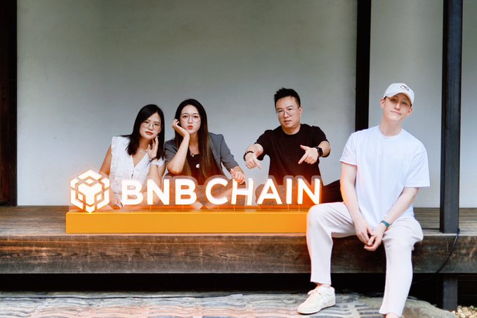 Four people pose in front of a glowing 'BNB CHAIN' sign, including a person in a white outfit and cap seated on the right.