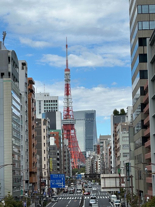 Tokyo Tower, an iconic orange-and-white communications tower, rises prominently above a bustling urban street lined with modern high-rises under a bright, partly cloudy sky—a perfect welcome to the city for an ETHGlobal event.