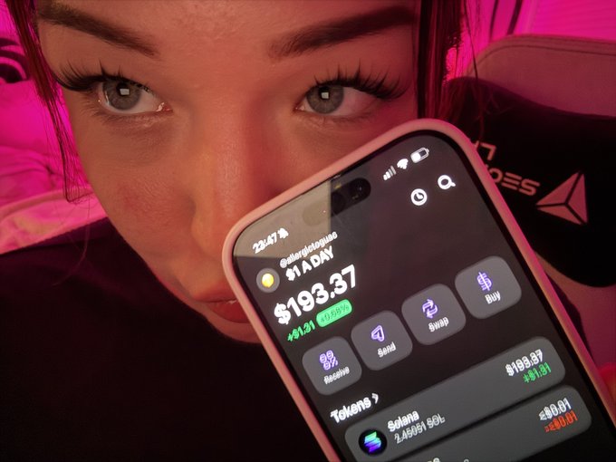 A close-up selfie of a young woman with a smartphone covering the lower half of her face, displaying a crypto app balance of $193.37, with the token price at $193.97 (+$1.91) and Solana at 2,45451 SOL, $0.01 (+$0.01), bathed in pink lighting.