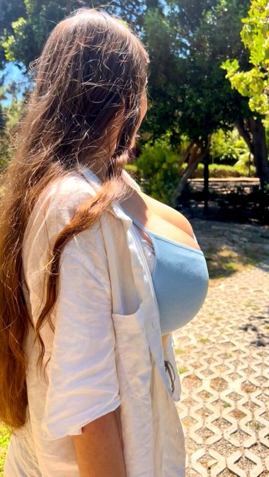 A woman with long wavy brown hair wearing a white shirt over a light blue tube top stands outdoors in a sunlit area.