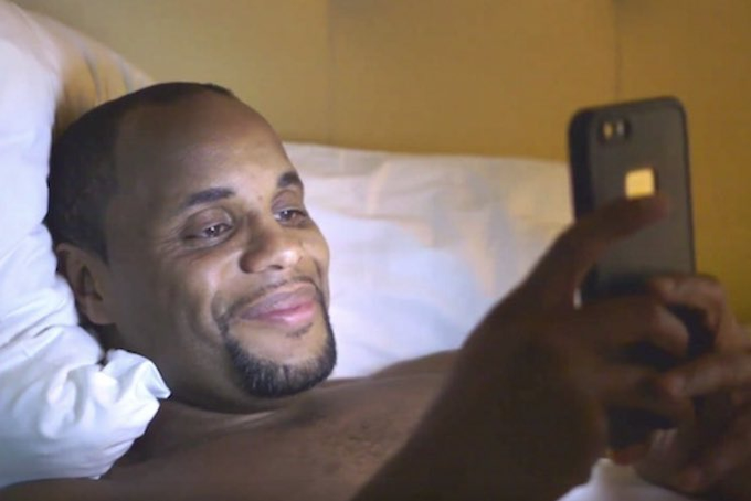 A man with a goatee lies in bed, smiling contentedly as he looks at his smartphone, appearing to be enjoying a moment of leisure with the phone's flash visible.