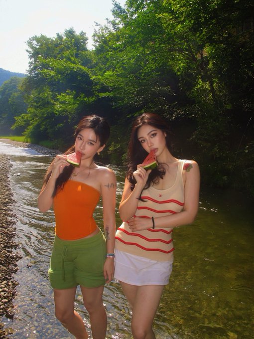 Two young women stand by a rocky stream in a wooded area, both eating slices of watermelon; the woman on the left wears an orange tube top and green shorts, while the woman on the right wears a striped tank top and white shorts.