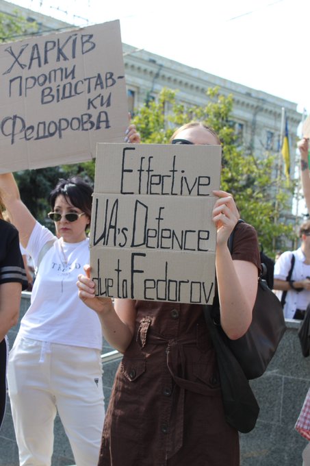 A protest scene where demonstrators hold cardboard signs; one sign in Ukrainian reads 'ХАРКІВ ПРОТИ ВІДСТАВКИ ФЕДОРОВА' (Kharkiv against Fedorov's resignation) and another in English reads 'Effective UAVs Defence due to Fedorov'.