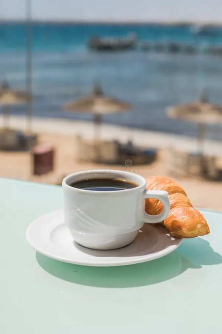 A cup of coffee sits on top of a white saucer with an orange pastry that looks like croissant toast inside. The background shows several beach umbrellas and chairs, and the water can be seen far off beyond them. There is also text in the image that reads "Happy new month hope we see a green chart".