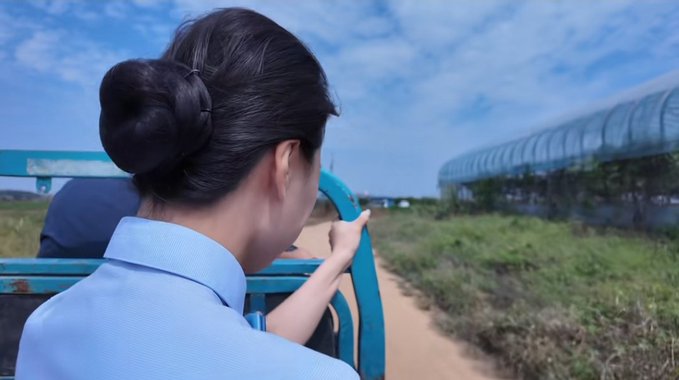 A young woman with her hair in a bun, wearing a light blue shirt, rides in an open vehicle on a dirt path, pointing toward a greenhouse structure in the distance.