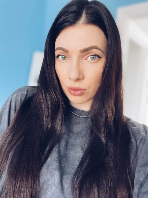 A close-up portrait of a young woman with long dark hair, wearing a gray shirt and a delicate silver necklace, looking directly at the camera against a light blue background.