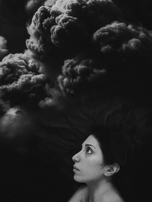 A black-and-white surreal portrait of a woman looking upward, with a massive, dramatic dark cloud formation rising from her head like thoughts or smoke against a black sky.