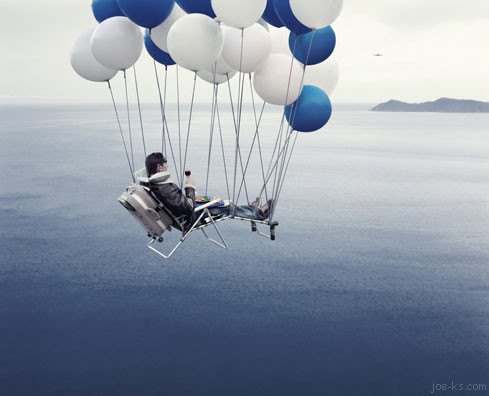 A surreal image shows a man with glasses and short blonde hair, smiling broadly while suspended in mid-air by a cluster of white balloons, drifting over what appears to be a vast landscape. The image, credited to "joe-ks.com" in the corner, evokes a whimsical, carefree mood that aligns with the playful, lighthearted spirit of the accompanying post's casual, cryptic message.