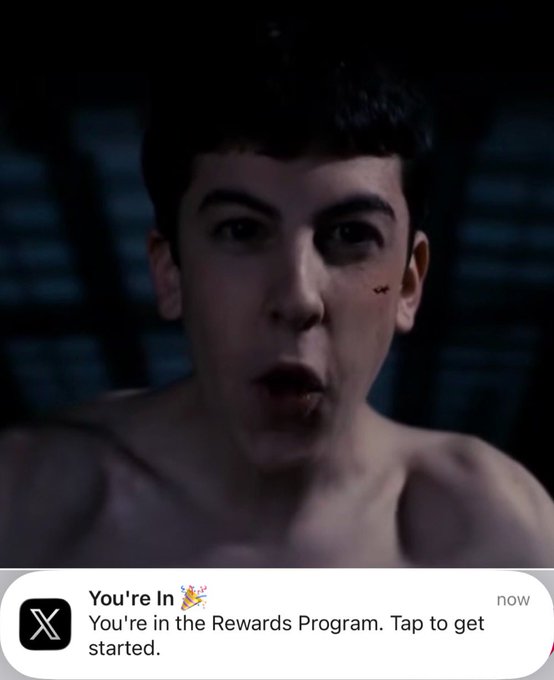 A close-up shows a shirtless young man with dark hair in a dimly lit setting, with an X (Twitter) notification at the bottom reading 'You're In 🎉 You're in the Rewards Program. Tap to get started.'