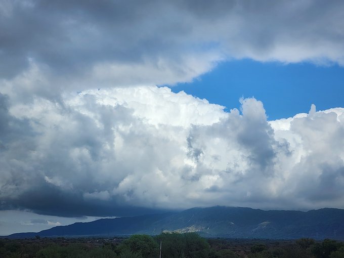A dramatic landscape features towering cumulus clouds billowing over a distant mountain range, with a thin strip of green vegetation in the foreground beneath a partly cloudy sky. The image's sweeping natural scenery complements the post's enthusiastic "Looks great" comment, suggesting the poster is admiring this stunning sky view.