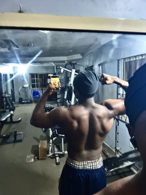 A shirtless man in a gym takes a mirror selfie with his phone while flexing his back muscles, wearing a dark cap and blue shorts with a checkered waistband.
