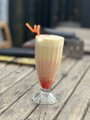 A small shot glass filled with a layered cream and red beverage, garnished with a small orange twist, sitting on a weathered wooden surface.