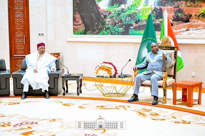 Two men sit in an ornate room for a diplomatic meeting; the man on the left wears traditional white robes and a red fez, while the man on the right wears a grey suit, seated between the African Union and Indian flags.