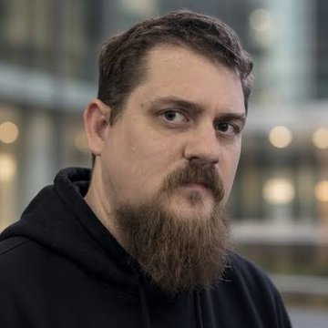 A portrait photo of a bearded man wearing a black hoodie, standing indoors with a blurred background.
