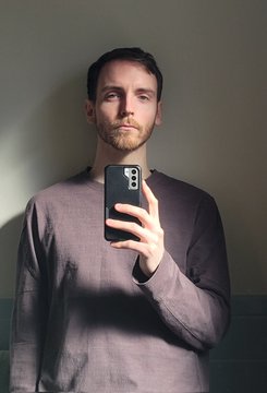 A selfie photo of a light-skinned man with a reddish beard wearing a purple long-sleeved shirt, holding a dark smartphone in front of a tiled wall.