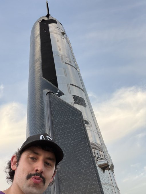 A man takes a selfie in front of SpaceX's towering Starship rocket, with his cap displaying the "SpaceX" logo. The image captures the impressive scale of the spacecraft ahead of its anticipated launch.