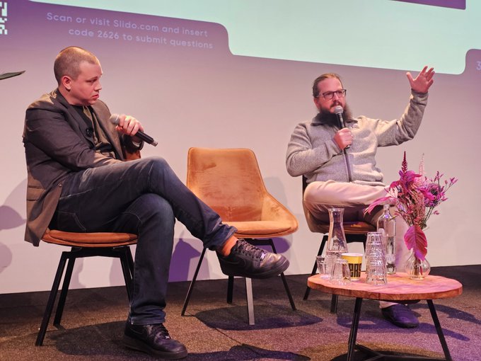 Two men seated on a stage during a panel discussion, with a slide reading 'Scan or visit Slido.com and insert code 2626 to submit questions', a flower arrangement, and water bottles on a small table beside them.