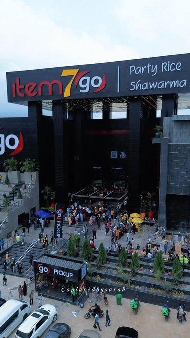 An aerial view of a large, modern 'item 7 go' store advertising 'Party Rice' and 'Shawarma', with crowds of people gathered outside near pickup stations.