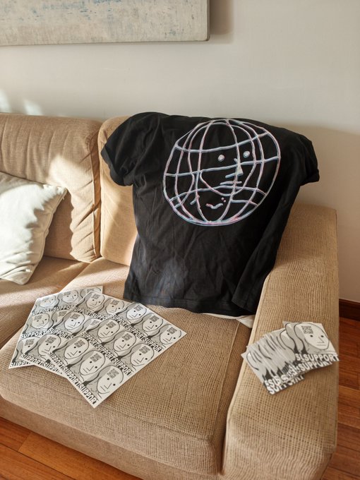 A black t-shirt featuring a neon-style globe smiley face design rests on a tan corduroy couch, accompanied by sheets of matching stickers scattered on the cushions.