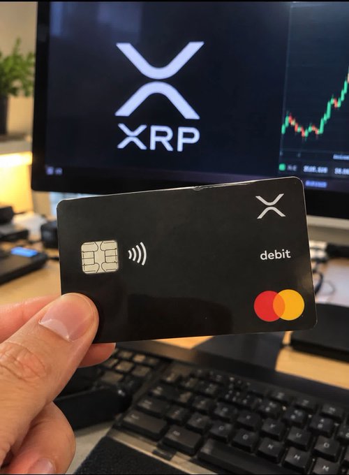 A hand holding a black XRP debit card in front of a computer monitor displaying the XRP logo and a cryptocurrency trading chart.