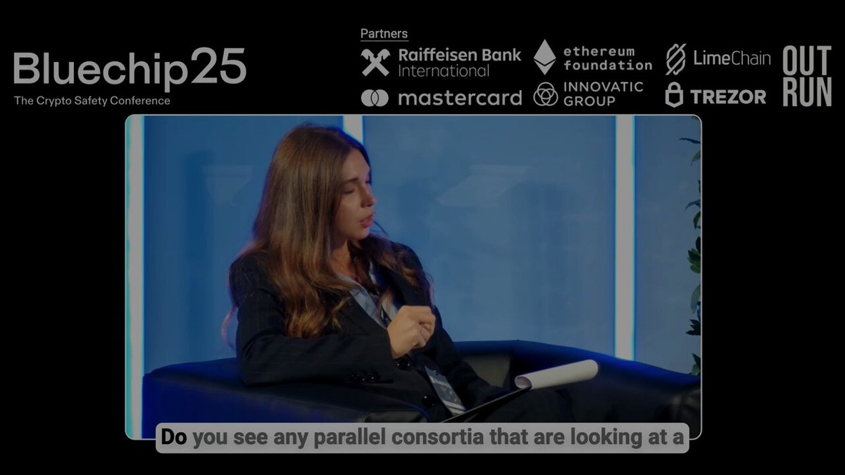 The image shows a woman sitting in an office chair, wearing a black suit jacket with her hands resting on her lap. The background of the image features a blue wall with various logos displayed on it, including "Bluechip25", "EUR stablecoins won'nt be winner-take-all. There' s room for two or three issuers. That's what Jan Sell is betting as the CEO of @QivalisEU , a group of banks taking on the current leader: @Circle 's EURC." and "@BKronfellner from BCG explain at Bluechip25". The woman appears to be engaged in a conversation or presentation related to these logos.