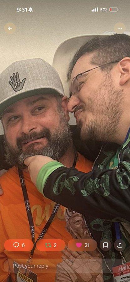 A screenshot from a social media app showing two men at what appears to be a gaming convention, one wearing an orange shirt and grey cap, with engagement metrics visible (6 comments, 2 retweets, 21 likes).
