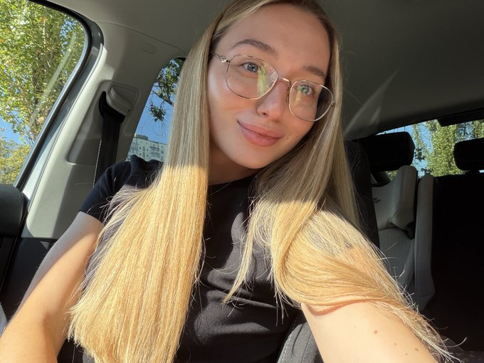 A young woman with long blonde hair and glasses takes a selfie inside a car, with sunlight streaming through the window. The casual, approachable image complements her introduction as a 24-year-old content creator and mom open to connecting with new founders and builders.
