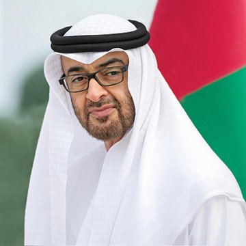 A portrait photograph of a man wearing a traditional white Emirati headdress (ghutra) with a black agal, glasses, and a white kandura, posed against a red and green background resembling the UAE flag.