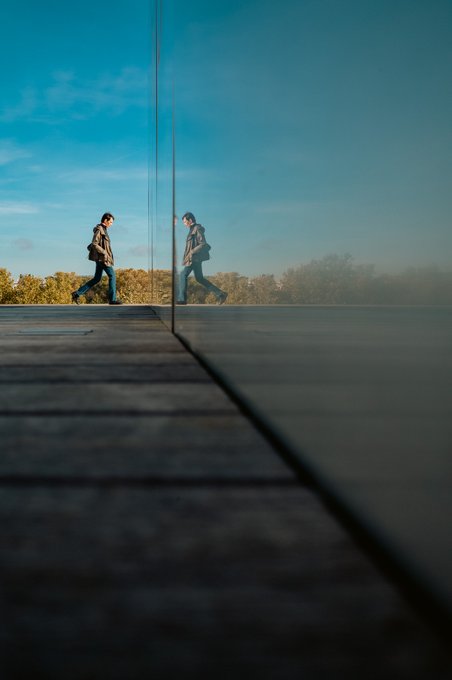 An artistic photo of a person walking along a road, with their reflection visible in a glass building, creating a mirrored composition against a blue sky.
