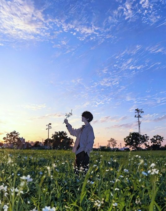 A young man in a beige jacket standing in a field of white flowers at sunset, holding up a small bouquet against a vast blue sky.