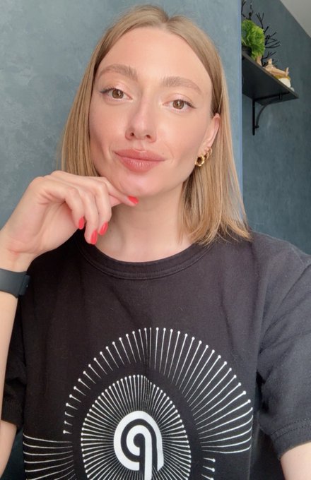 The image features a woman wearing a black t-shirt with a white design of a sunburst shape. She has blonde hair and is posing for the camera while looking at the camera, giving off a confident and stylish vibe. The background appears to be a wall or a gray surface, which complements her outfit and overall appearance.