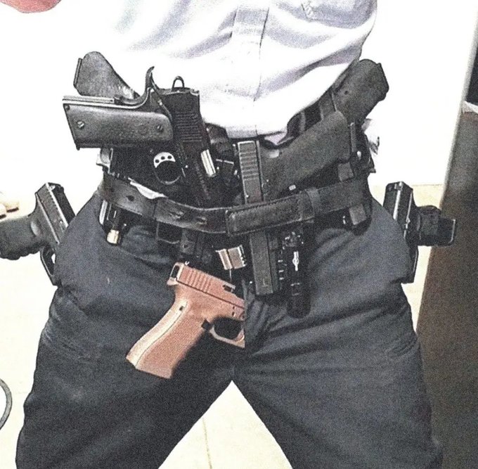 A person is shown from the torso down wearing dark pants and a belt laden with multiple holstered handguns, including a semi-automatic pistol with a brown grip prominently displayed in the front center. Additional firearms are holstered on both hips and at the waist, suggesting an armed security or tactical context.