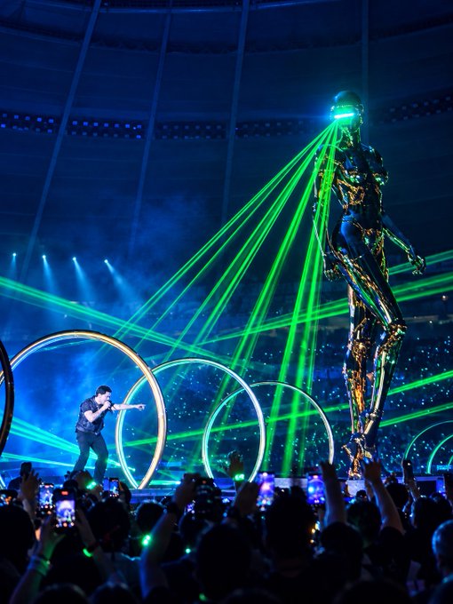 A large concert arena with a performer on stage, green laser beams, and a giant golden humanoid statue towering above the audience.